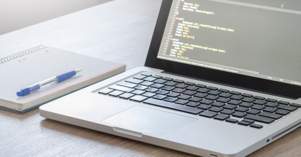 Laptop with code editor open, notebook and pen on wooden desk, perfect for tech and work themes.